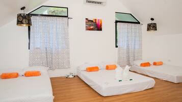 Une chambre d’hôtel avec deux lits simples, chacun doté de draps blancs et d’oreillers orange. On y trouve une petite table recouverte d’une nappe blanche, sur laquelle est posé un objet vert. La chambre a un plafond en pente avec un puits de lumière et un appareil de climatisation fixé au mur.