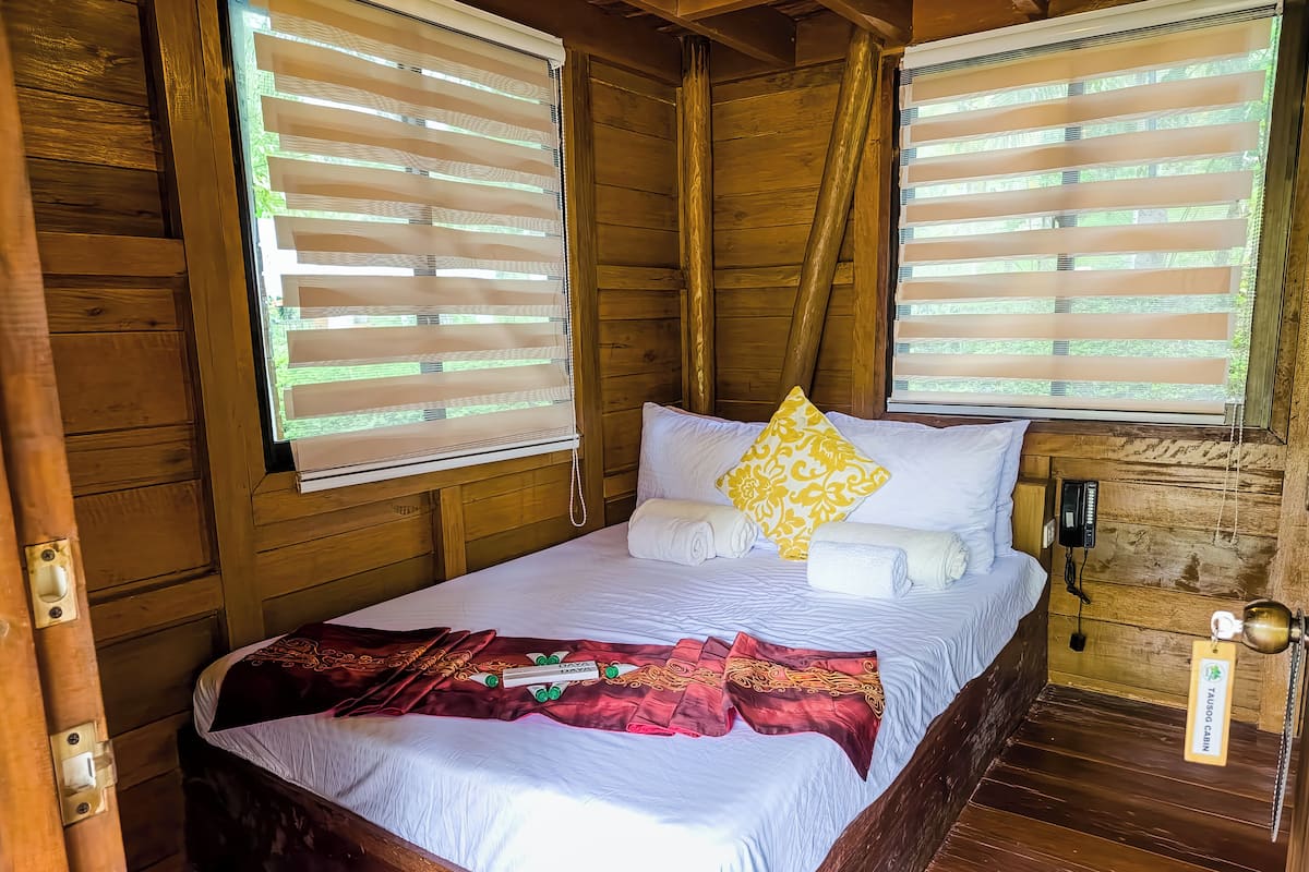 A wooden cabin room with a bed, two windows with blinds, a bedside table, and a lamp.
