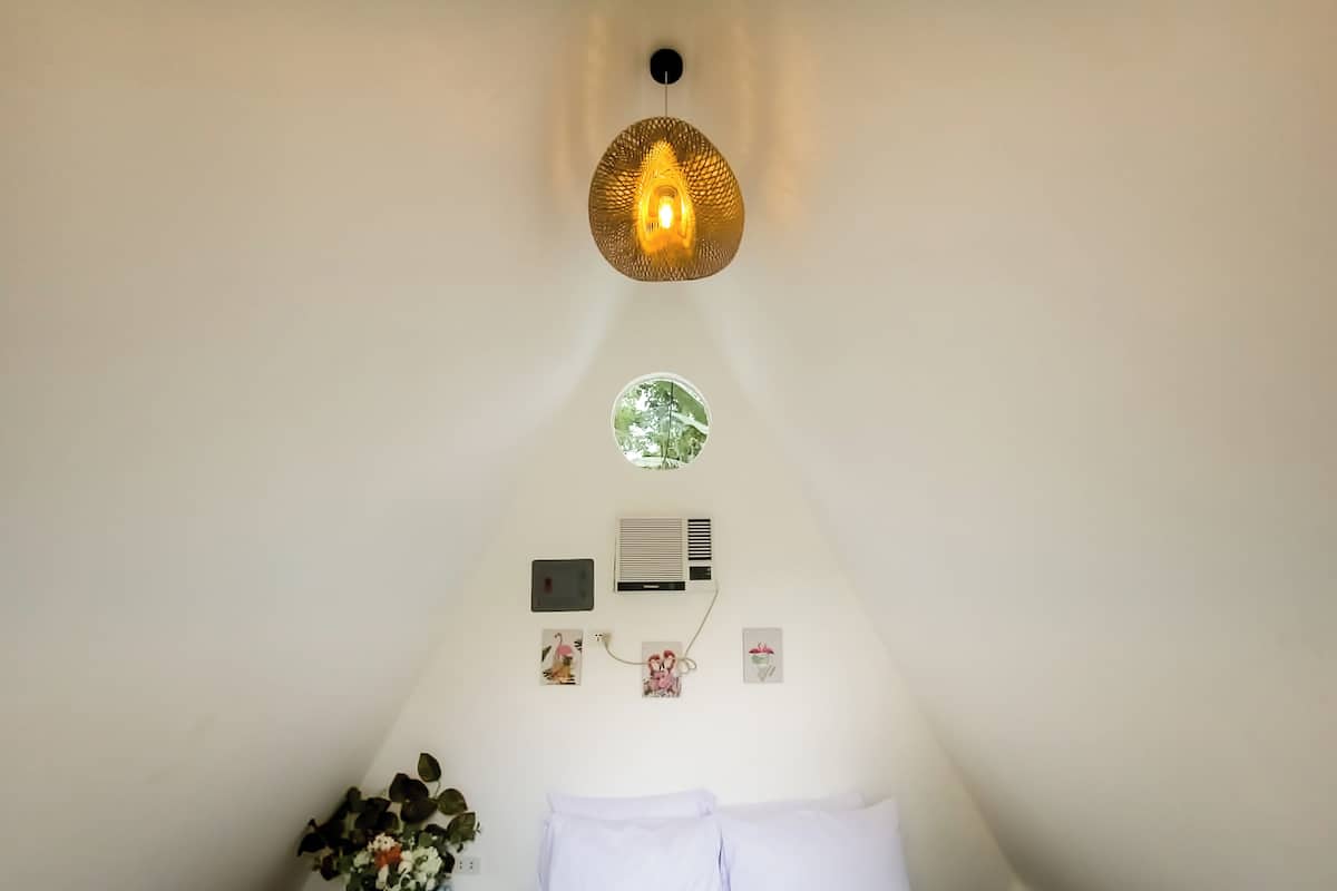 A bedroom with a bed, a nightstand with a plant, a wall-mounted air conditioner, and a unique circular light fixture.