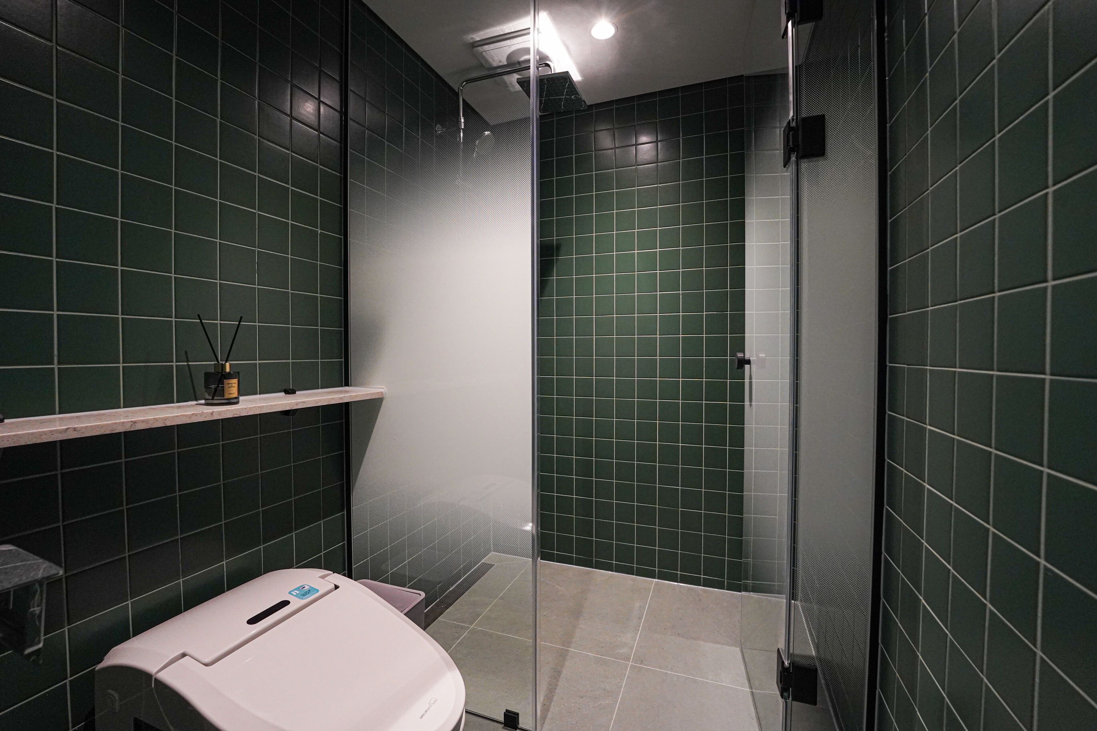 A modern bathroom with green tiles, a glass shower enclosure, and a white countertop with a soap dispenser.