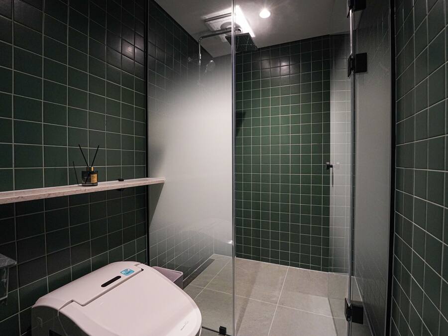 A modern bathroom with green tiles, a glass shower enclosure, and a white countertop with a soap dispenser.