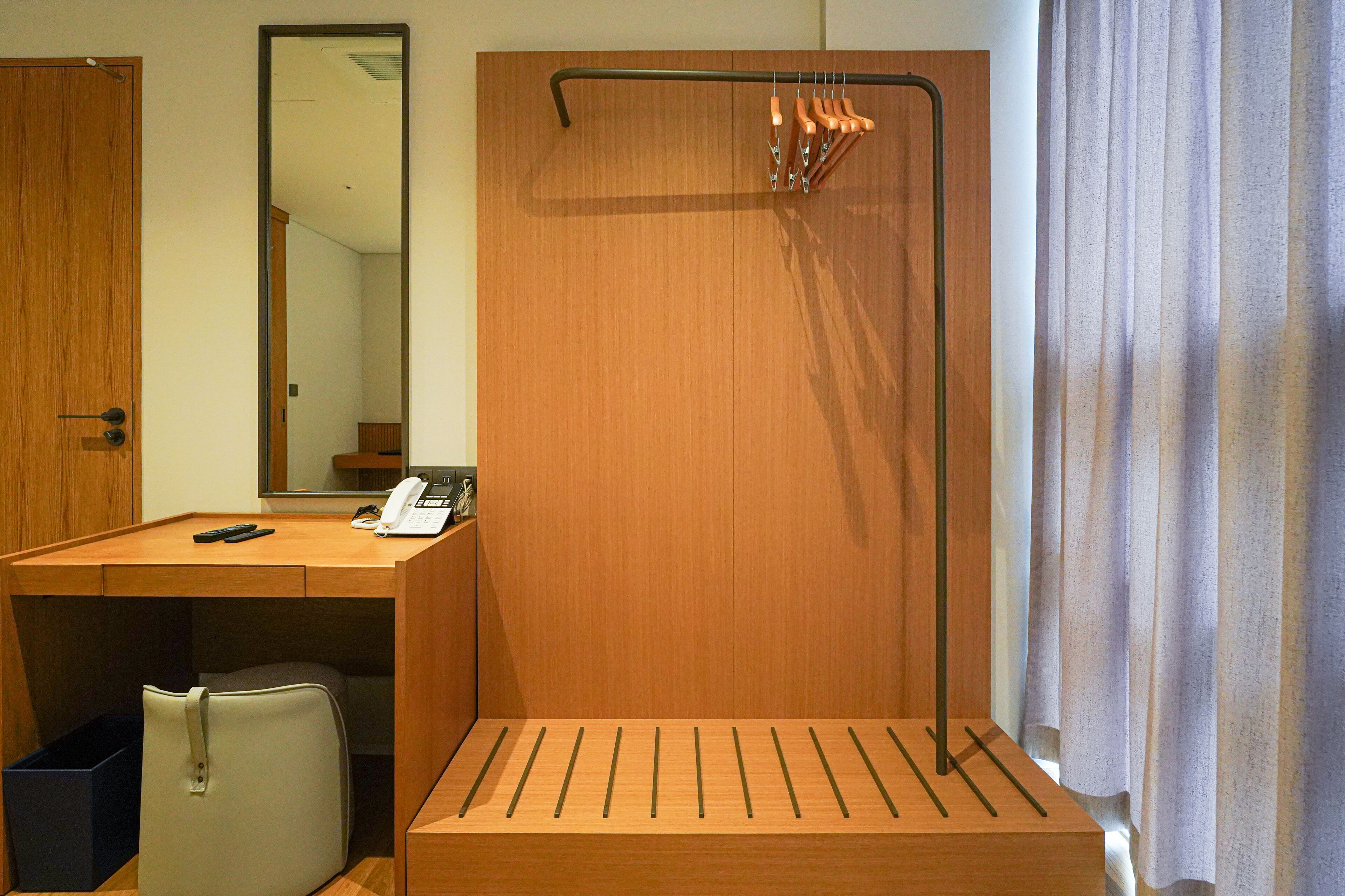 A hotel room with a wooden desk, a telephone, a mirror, and a hanging coat rack.