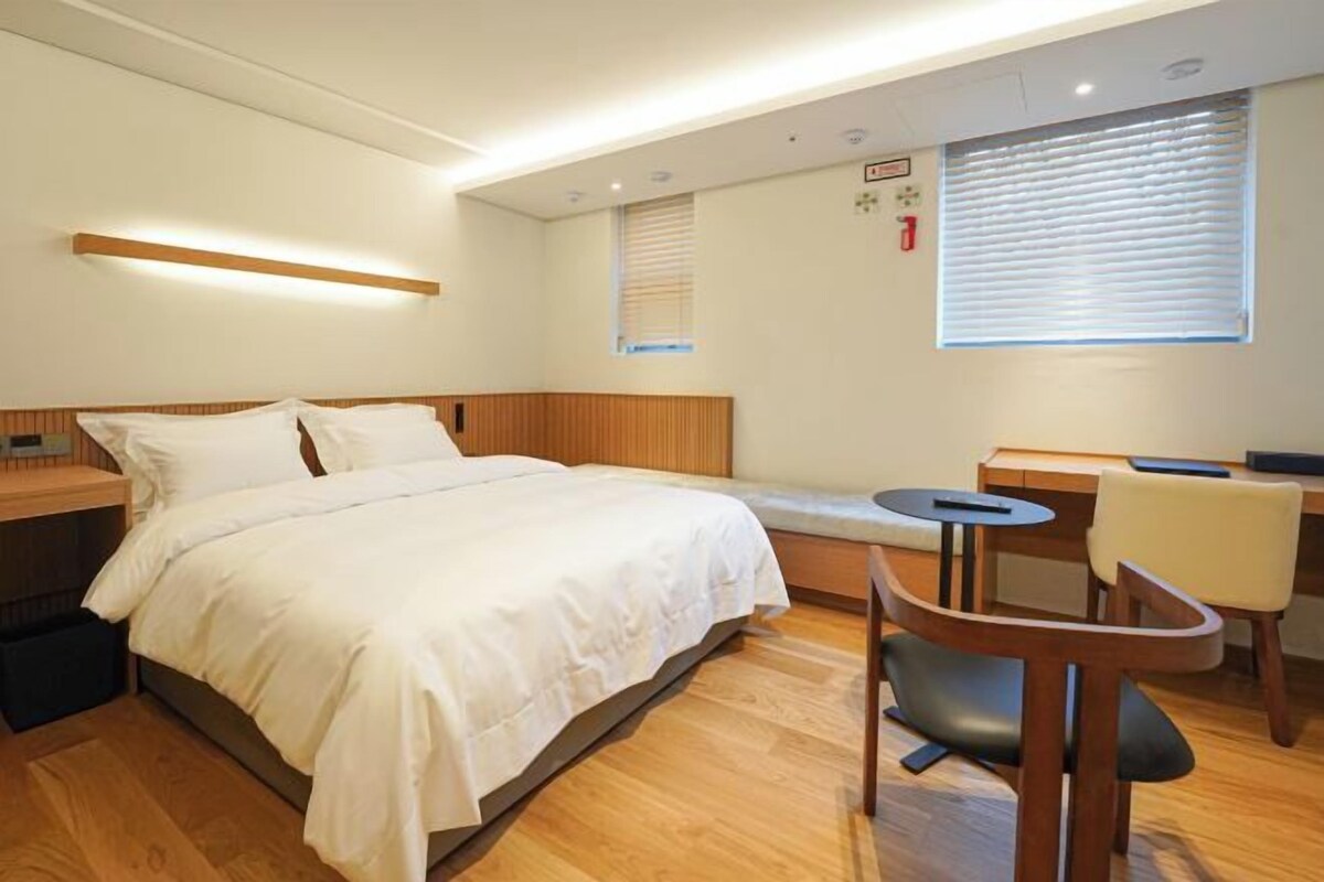 A bedroom with a bed, bedside table, chair, desk, and window.