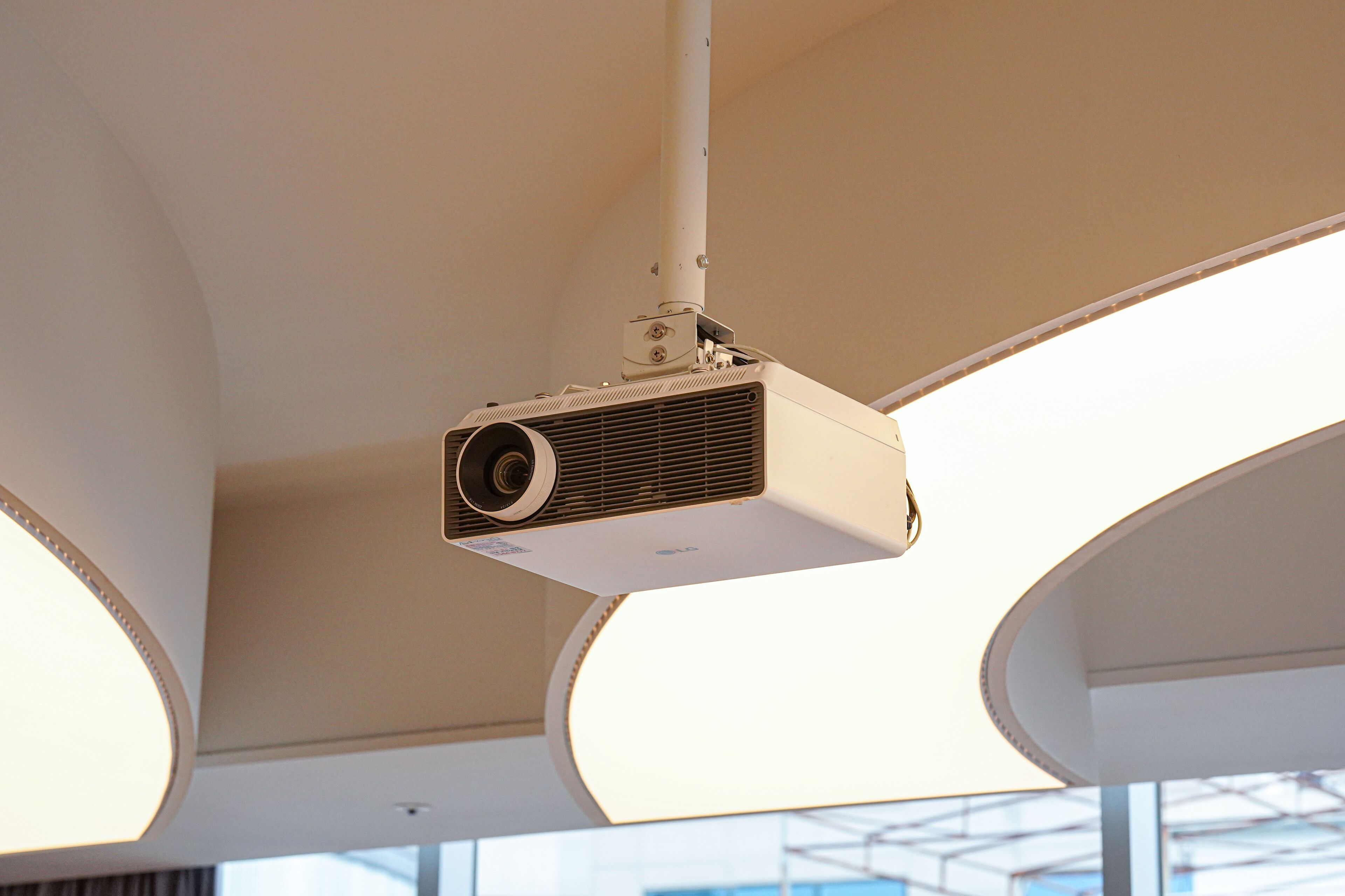 A ceiling-mounted projector in a modern room with curved ceiling panels and large windows.