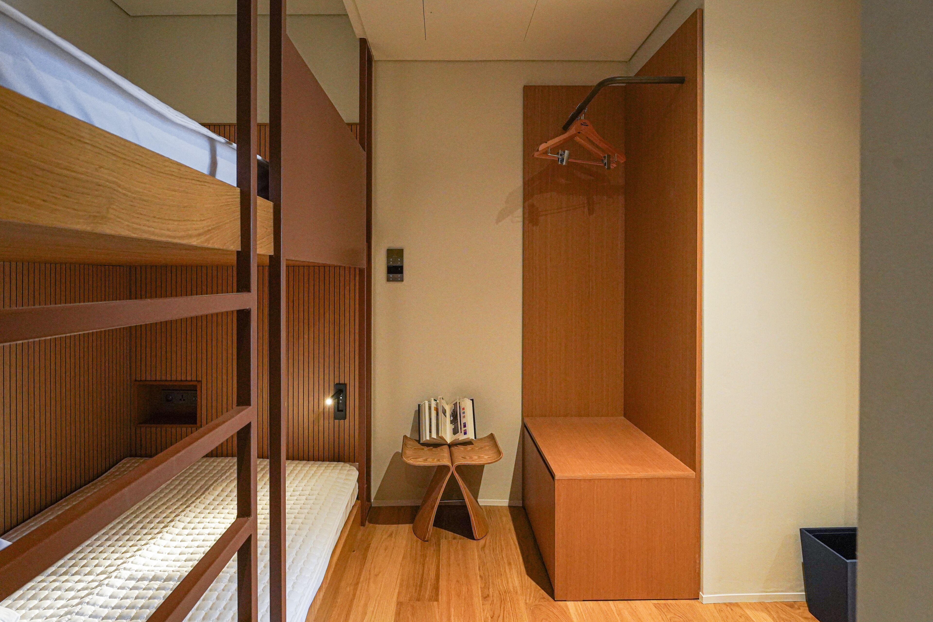A compact room with bunk beds, a small wooden table with books, a hanging coat rack, and a trash bin.