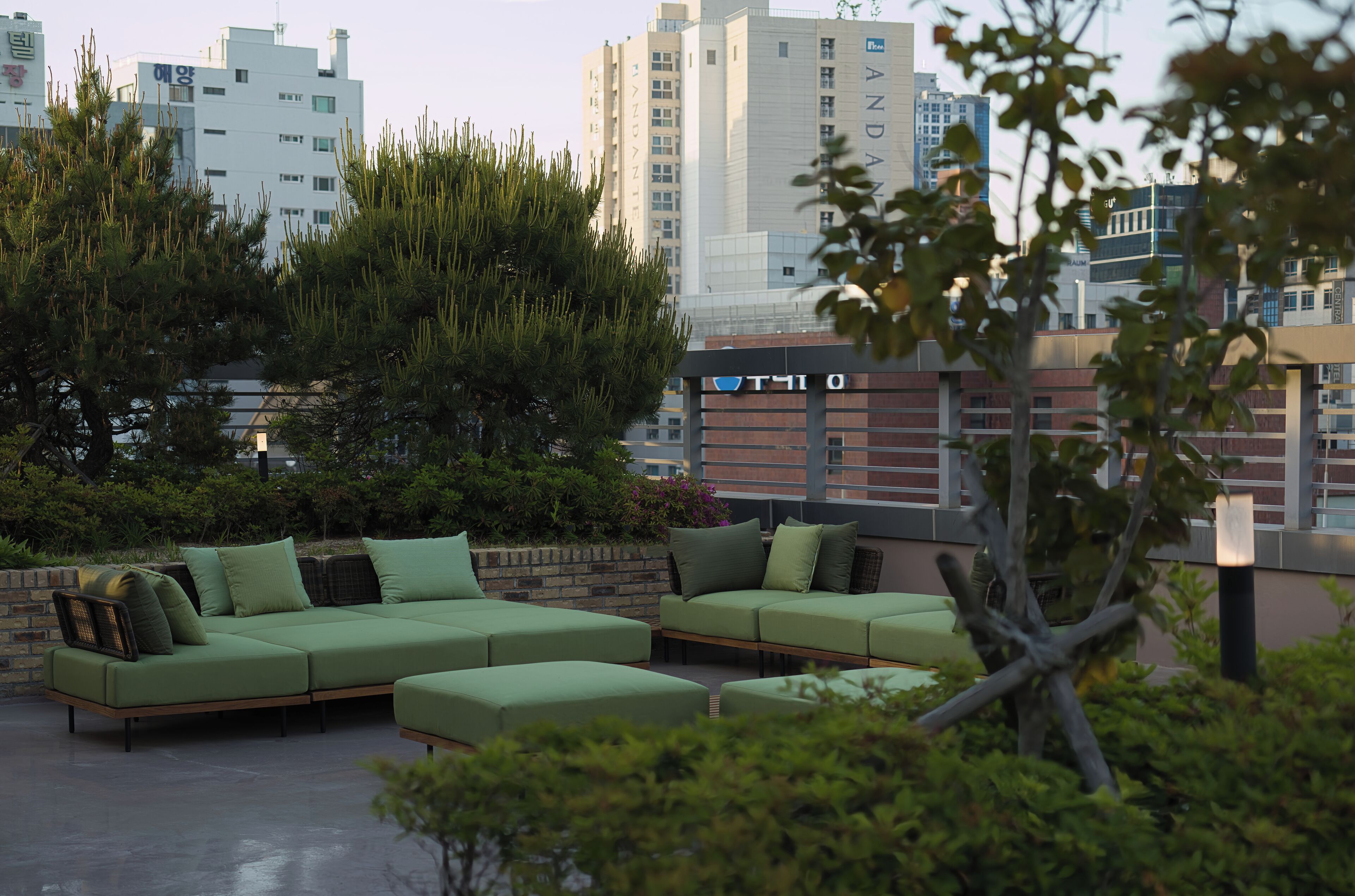 An outdoor seating area with green furniture, surrounded by greenery and a cityscape in the background.