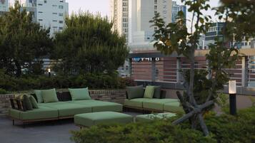 An outdoor seating area with green furniture, surrounded by greenery and a cityscape in the background.