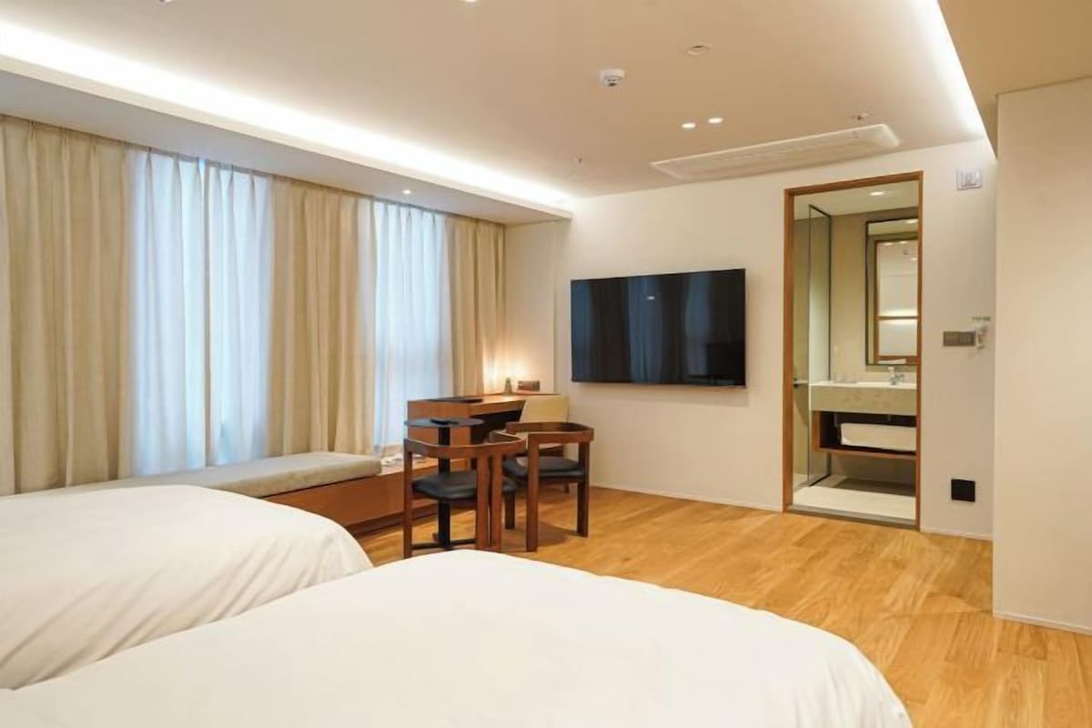 A hotel room with two beds, a desk with chairs, a TV, and a bathroom with a mirror.