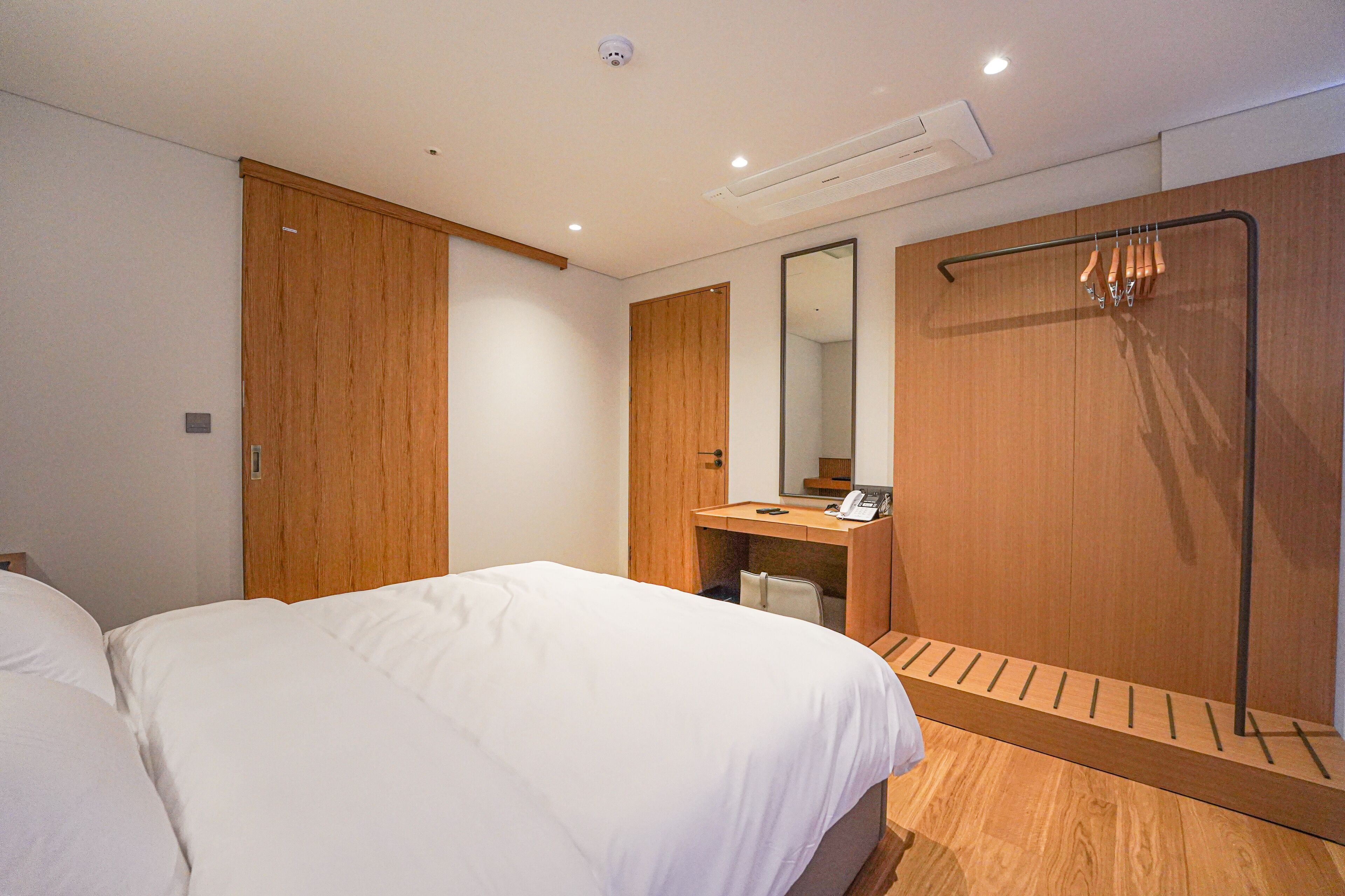 Een hotelkamer met een bed, een houten garderobe, een spiegel, een bureau met telefoon en een handdoekrek.