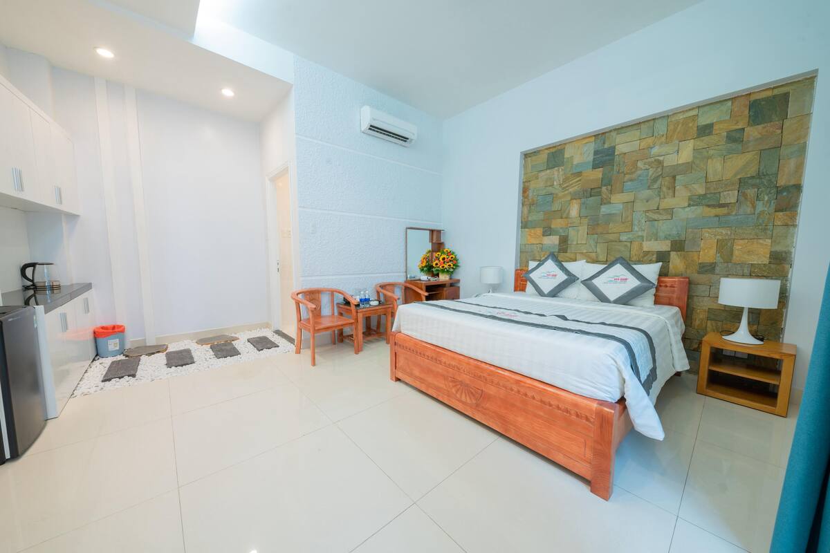 A hotel room with a wooden bed, a desk with a chair, a small table with a lamp, and a wall-mounted air conditioner.