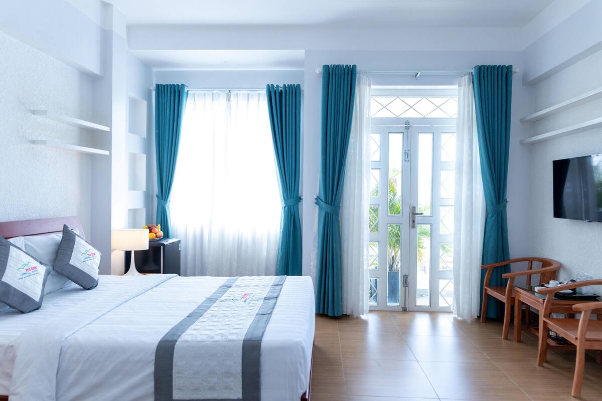 A hotel room with a bed, a desk, a chair, a TV, and a balcony with curtains.