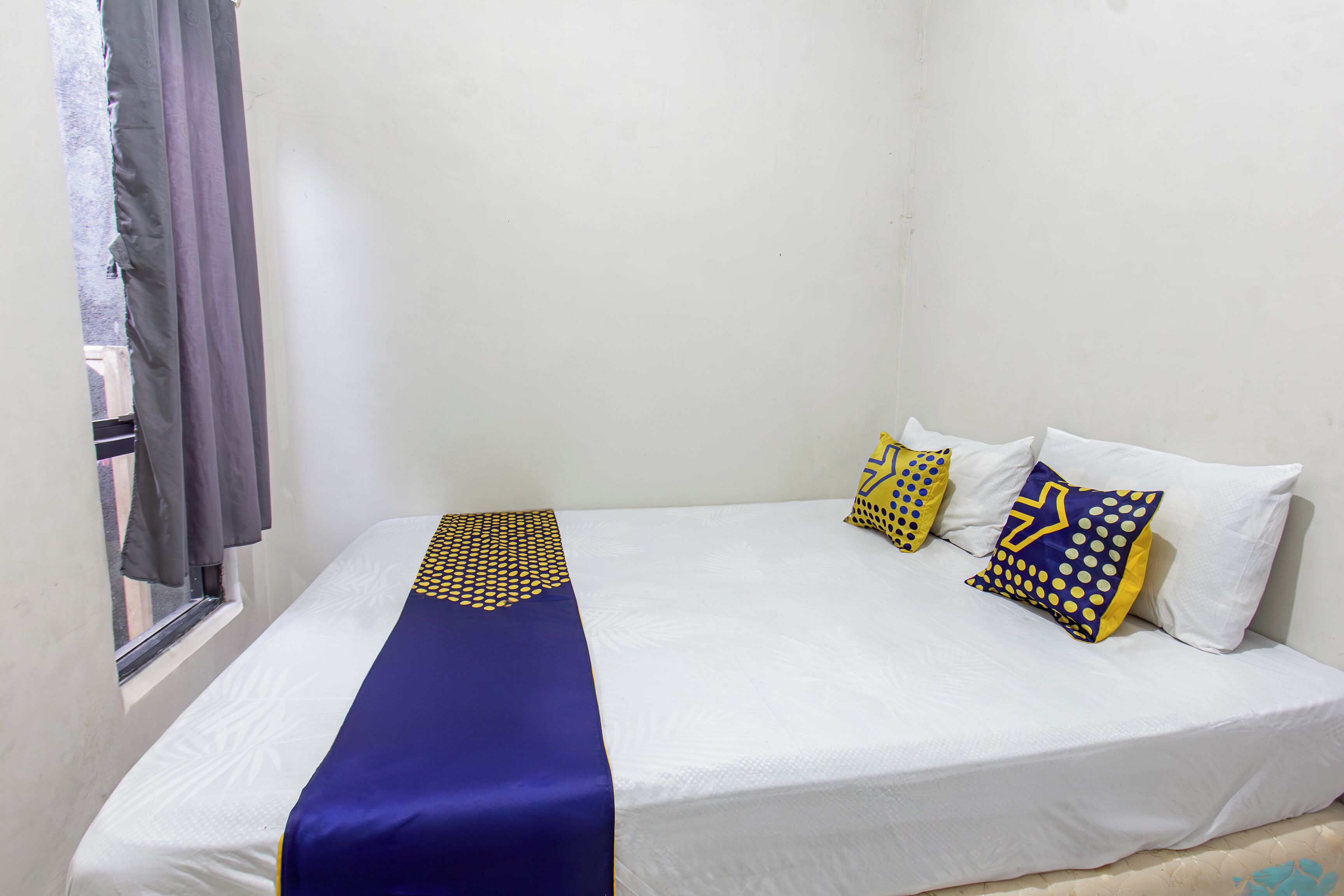 A single bed with a white mattress, a blue bedspread, and three decorative pillows with yellow and blue patterns.
