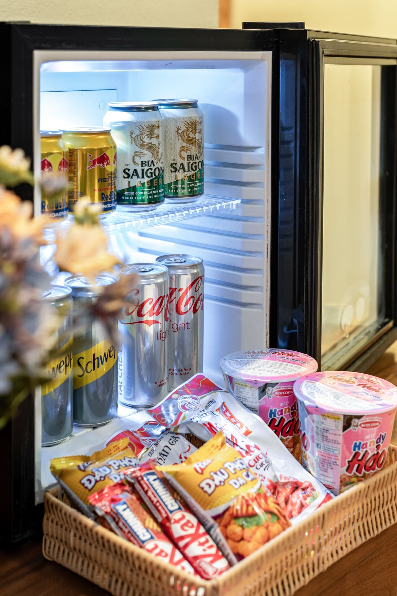 Un réfrigérateur rempli de boissons et d’en-cas, notamment des canettes de Bia Saigon Lager, de Coca-Cola Light, de Schweppes et divers produits d’épicerie emballés.