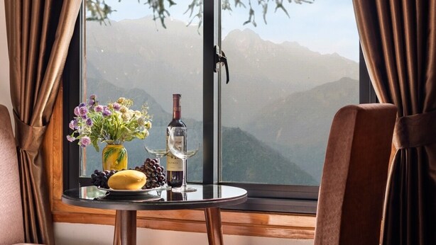 A room with a large window, two chairs, a table with a bottle and glasses, and a vase with flowers. - Tiger Sapa Hotel (Sa Pa)