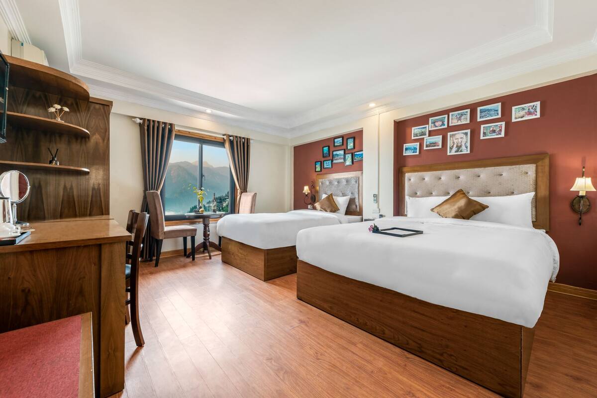 Une chambre d’hôtel avec deux lits, un bureau, une chaise, un miroir et une vue sur les montagnes par la fenêtre.