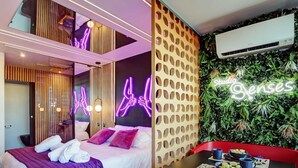 Una habitación de hotel moderna con una cama, un letrero de neón, un aire acondicionado instalado en la pared y un rincón de comedor con mesa y sillas.