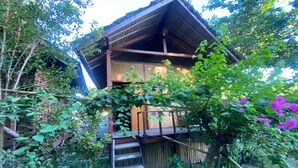 A rustic wooden structure with a thatched roof, surrounded by lush greenery and flowering plants. - SPOT ON 93511 Rinjana Homestay (Sembalun Lawang)