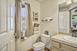 A bathroom with a toilet, sink, and a mirror. There are decorative wall art pieces and a sign that says "Please seat yourself." - Highland Retreat (Woodland Park)