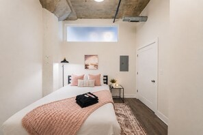 Un dormitorio con tragaluz, cama con ropa de cama rosa, mesita de noche con una planta y mesita auxiliar.