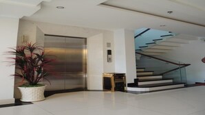 Um elevador moderno com acabamento metálico, um vaso de planta de folhas vermelhas e uma escadaria com guarda-corpo de vidro.