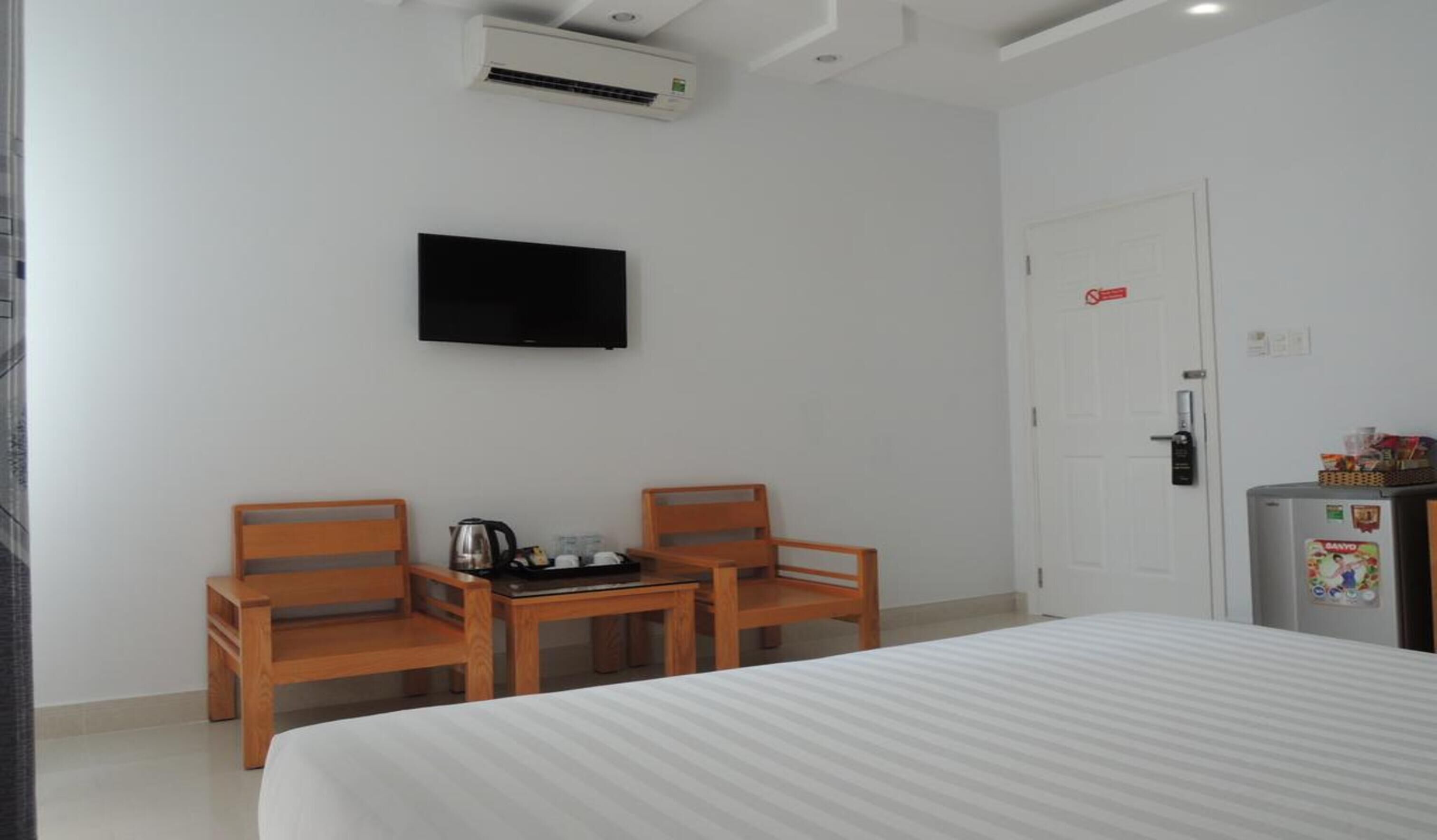 A hotel room with a bed, two wooden chairs, a small table with a kettle, a television on the wall, a refrigerator with magnets, and a door with a lock.