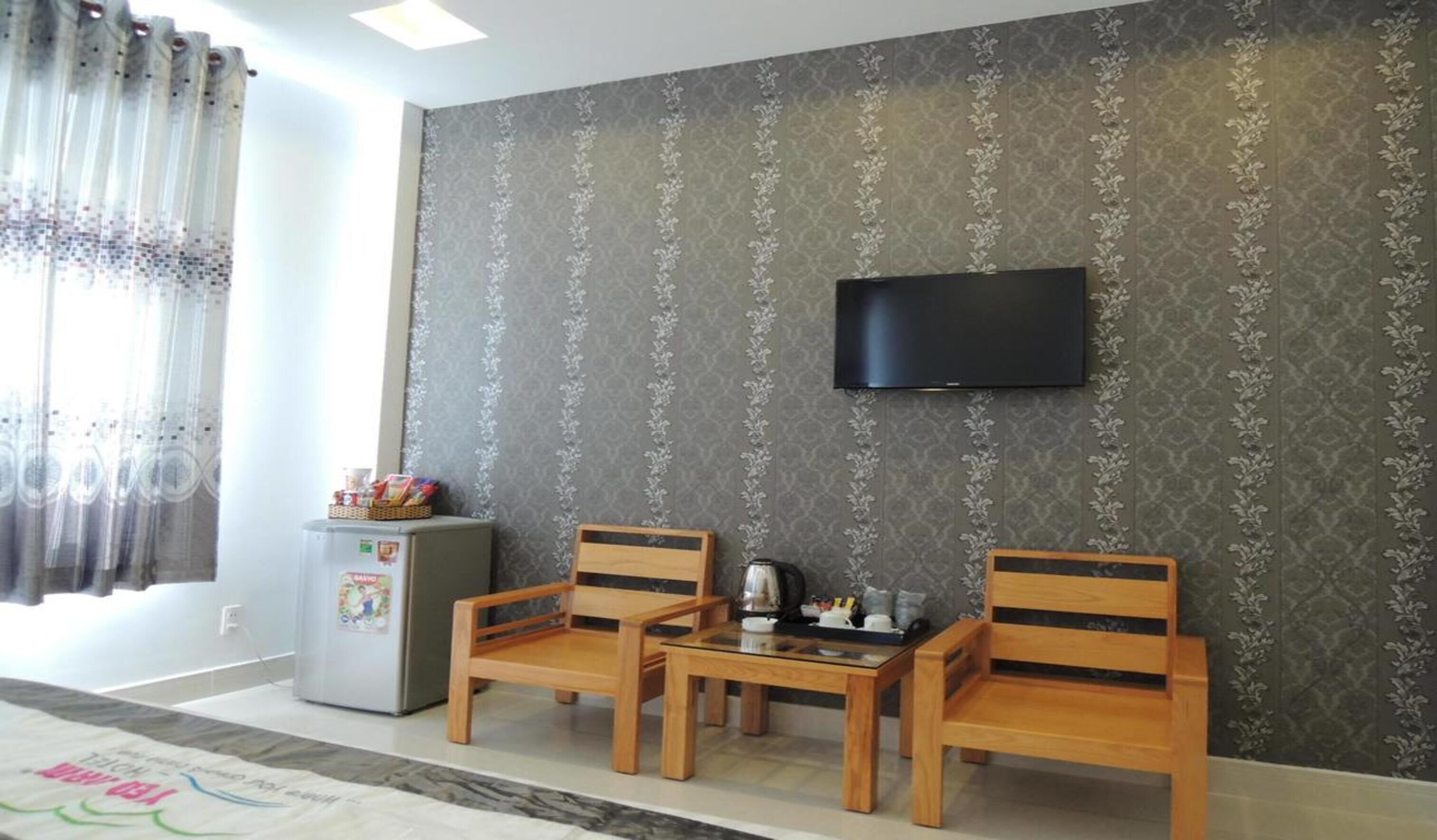 A living room with two wooden chairs, a small table, a television mounted on the wall, and a refrigerator with magnets.