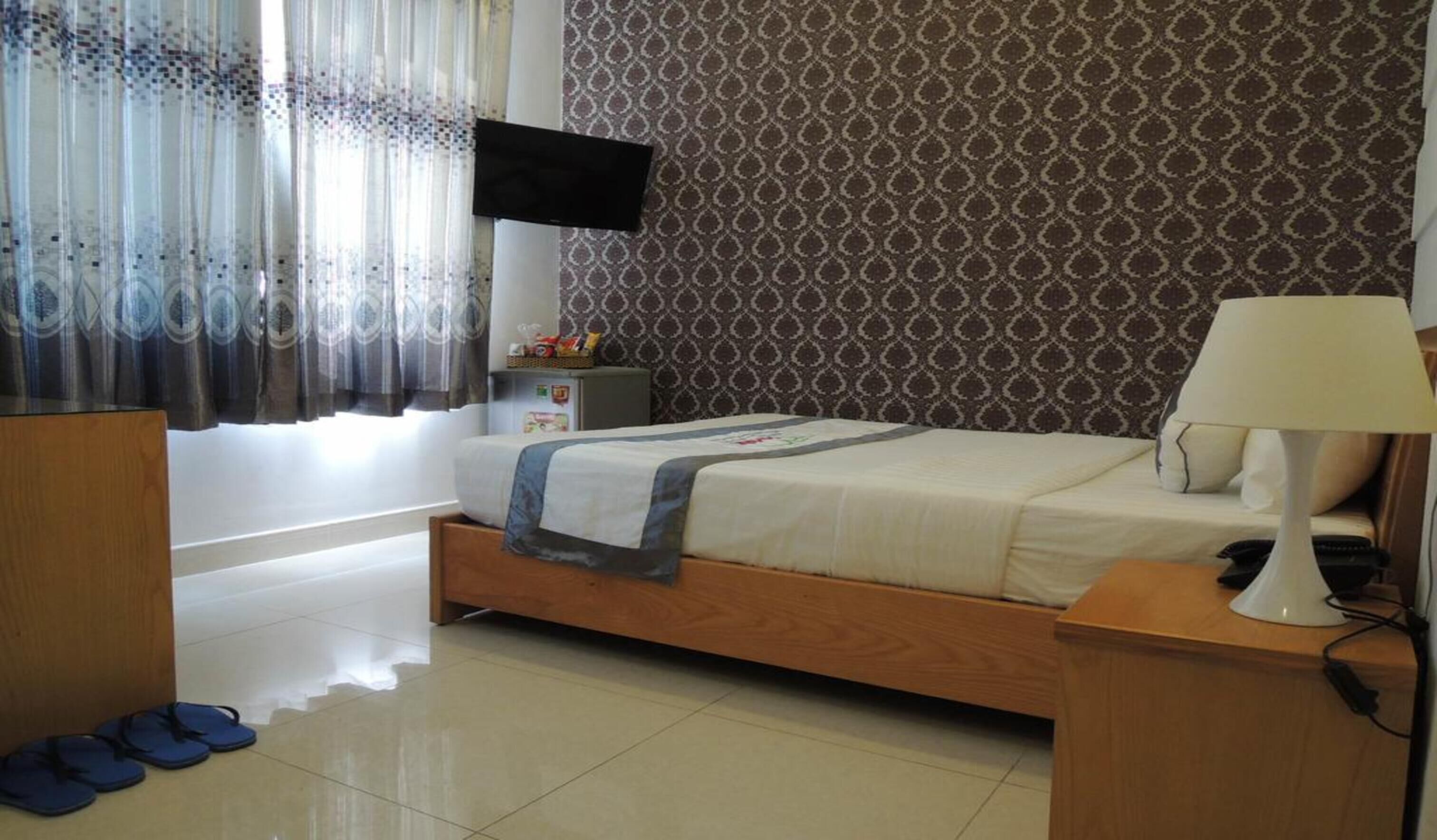 A bedroom with a bed, a TV mounted on the wall, a bedside table with a lamp, and a pair of slippers on the floor.