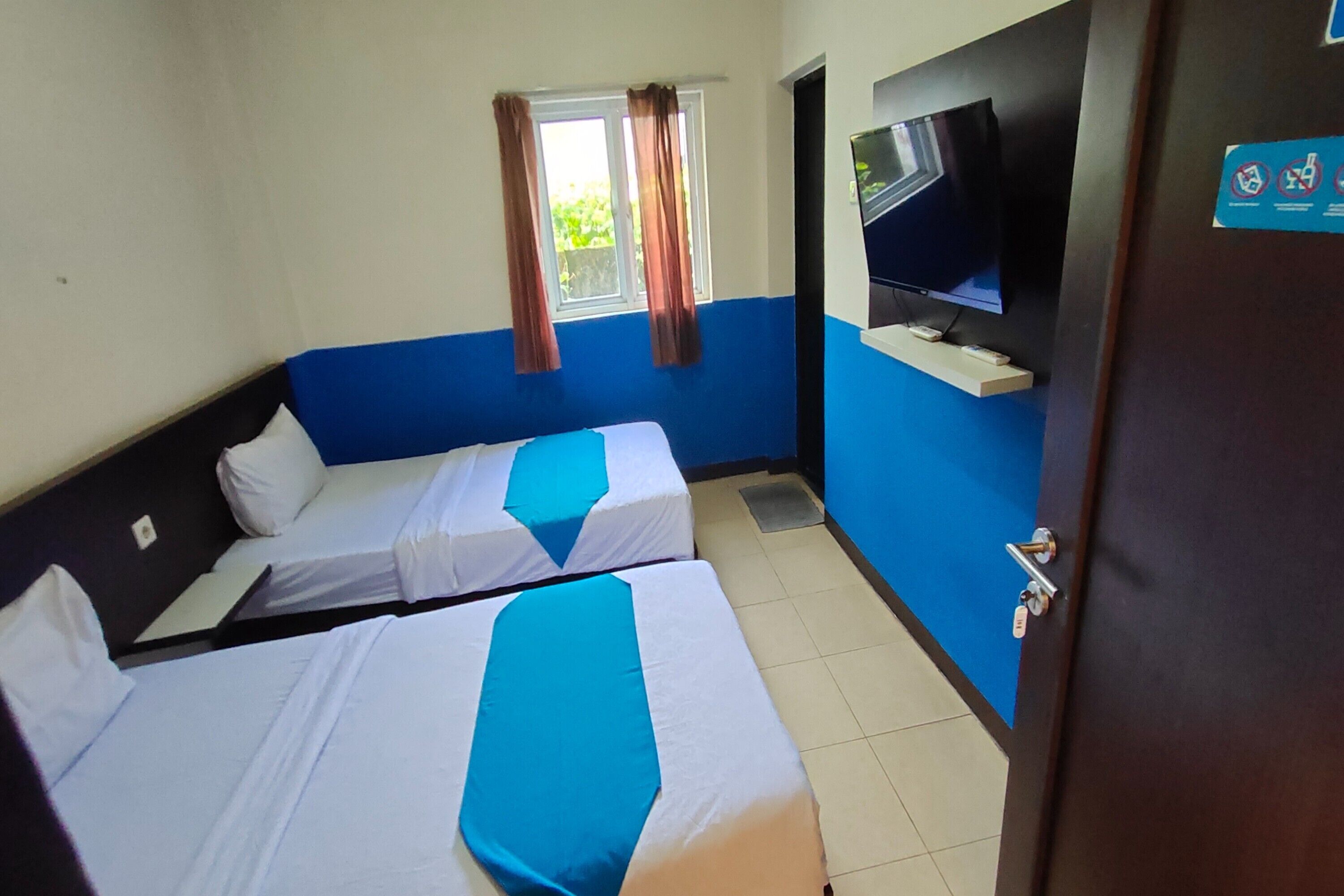 A hotel room with two single beds, a television mounted on the wall, and a window with curtains.