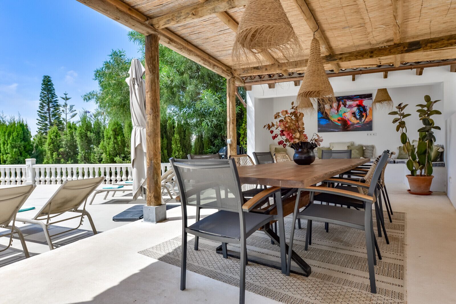 A covered outdoor dining area with a table and chairs, a potted plant, and a painting on the wall.