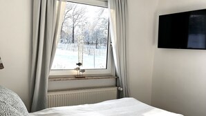 Un dormitorio con un ventanal grande, un radiador, una cama con colcha estampada, un televisor montado en la pared y una cesta de mimbre.