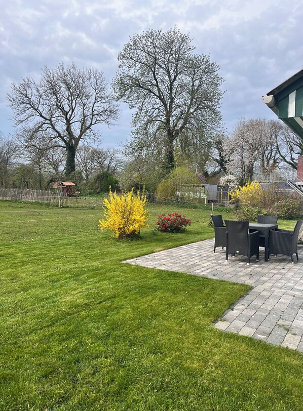 Ein gepflegter Rasen, eine Terrasse mit Gartenmöbeln und verschiedene Bäume und Sträucher.