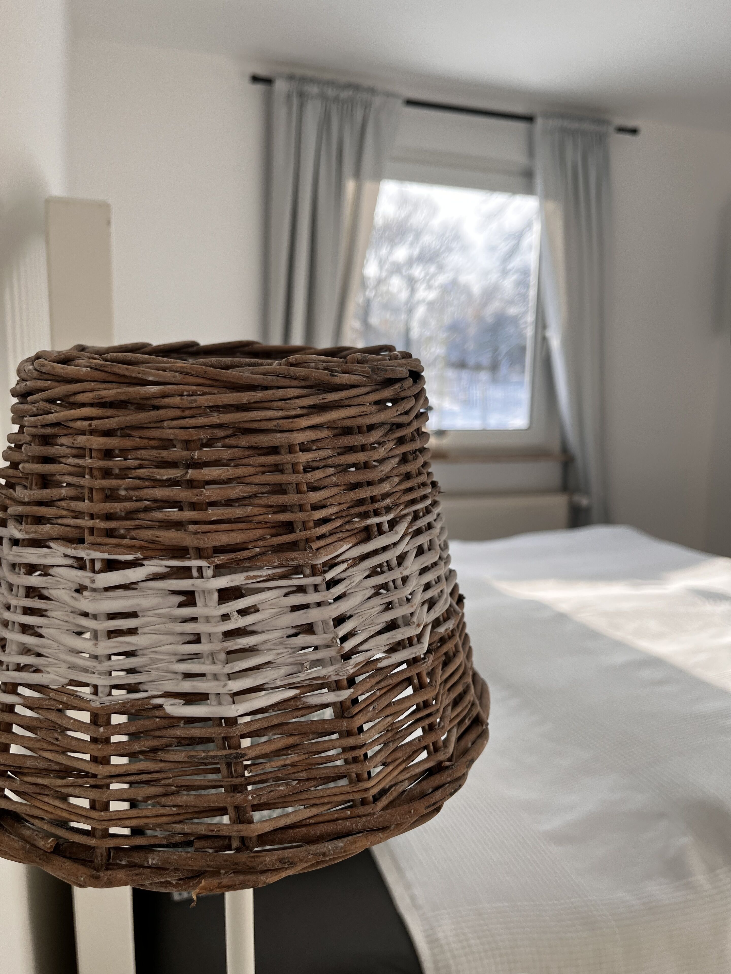A wicker lamp on a stand in a room with a bed and a window with curtains.
