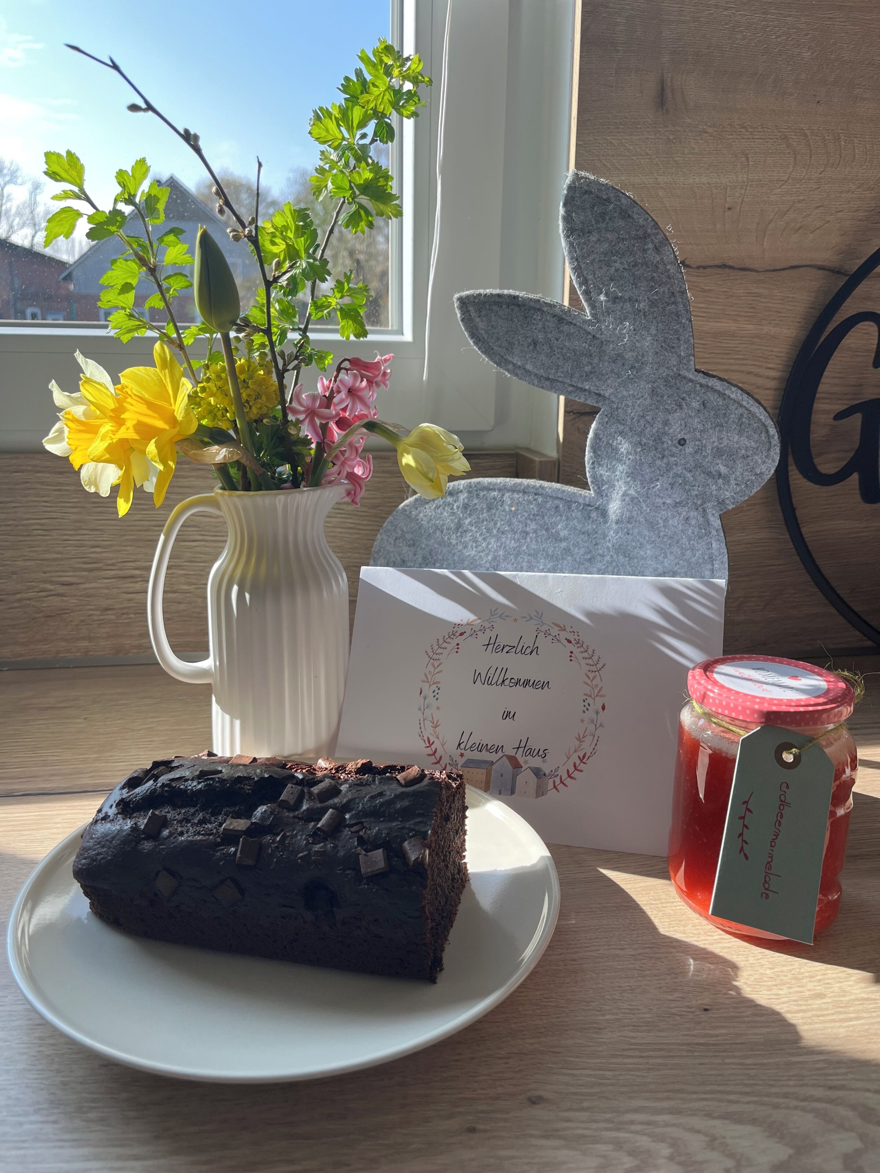 A slice of chocolate cake on a plate, a white vase with flowers, a greeting card, a felt bunny decoration, and a jar of jam.