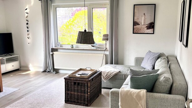 Una sala moderna con un sofá gris, una mesa de centro de mimbre, un televisor y una ventana con cortinas.
