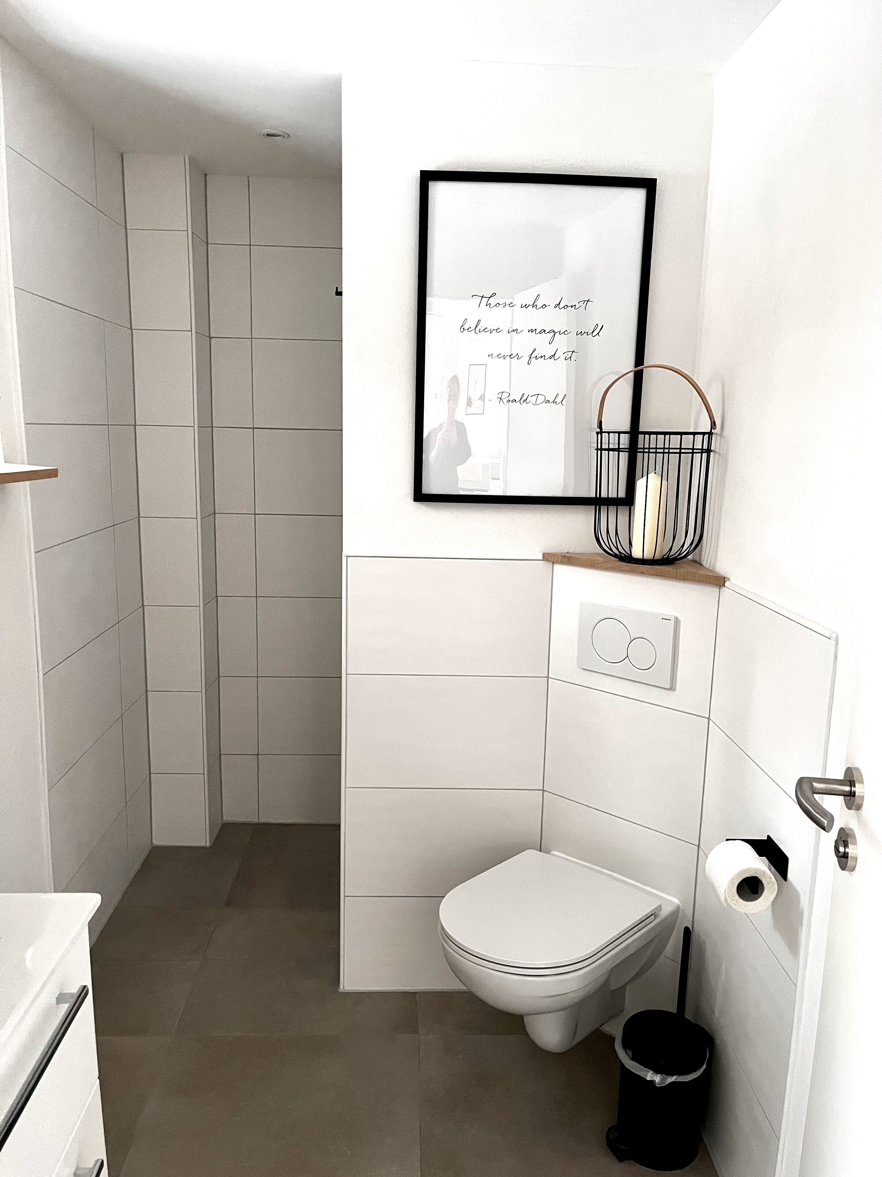 Un bagno moderno con piastrelle bianche, un water, un cestino nero e una citazione incorniciata al muro.