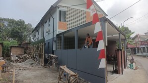 Un immeuble résidentiel en construction, avec un mur extérieur partiellement achevé, un ouvrier sur le toit et des matériaux de construction éparpillés aux alentours.