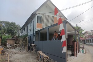A residential building under construction with a partially completed exterior wall, a worker on the roof, and construction materials scattered around.