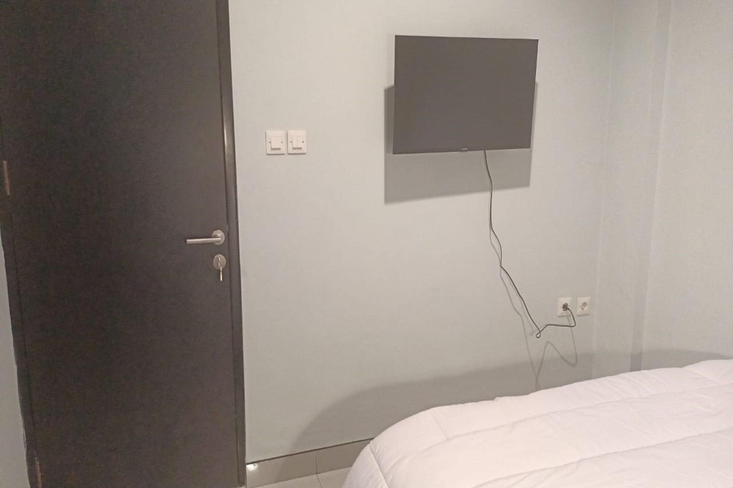 A bedroom with a bed, a door, a wall-mounted TV, and electrical outlets.