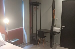 A hotel room with a bed, a desk with a chair, a small table, a wardrobe, a hanging rack, and a sink.