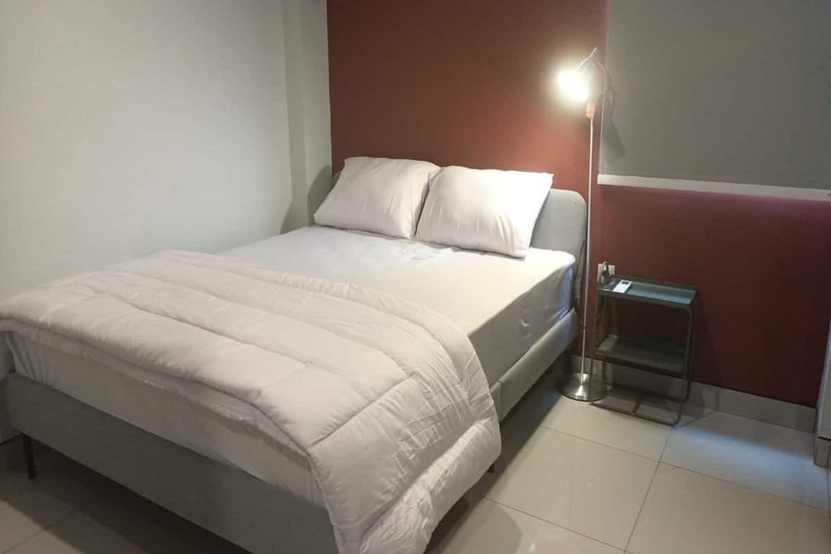 Een netjes opgemaakt bed met witte beddengoed en kussens, een nachtkastje met een lamp, en een rode accentmuur.