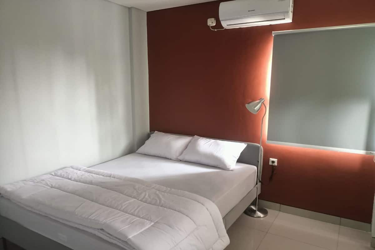 Een netjes opgemaakt bed met witte beddengoed in een kamer met een rode accentmuur, een witte airconditioning en een nachtkastje met een lamp.