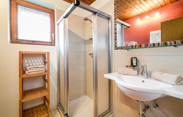 A bathroom with a glass-enclosed shower, a white sink, and a shelf with folded towels. - Apartments Tannheim (Tannheim)