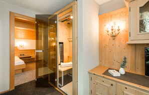 A modern bathroom with a glass shower enclosure, a wooden vanity, and a wall-mounted light fixture. - Apartments Tannheim (Tannheim)