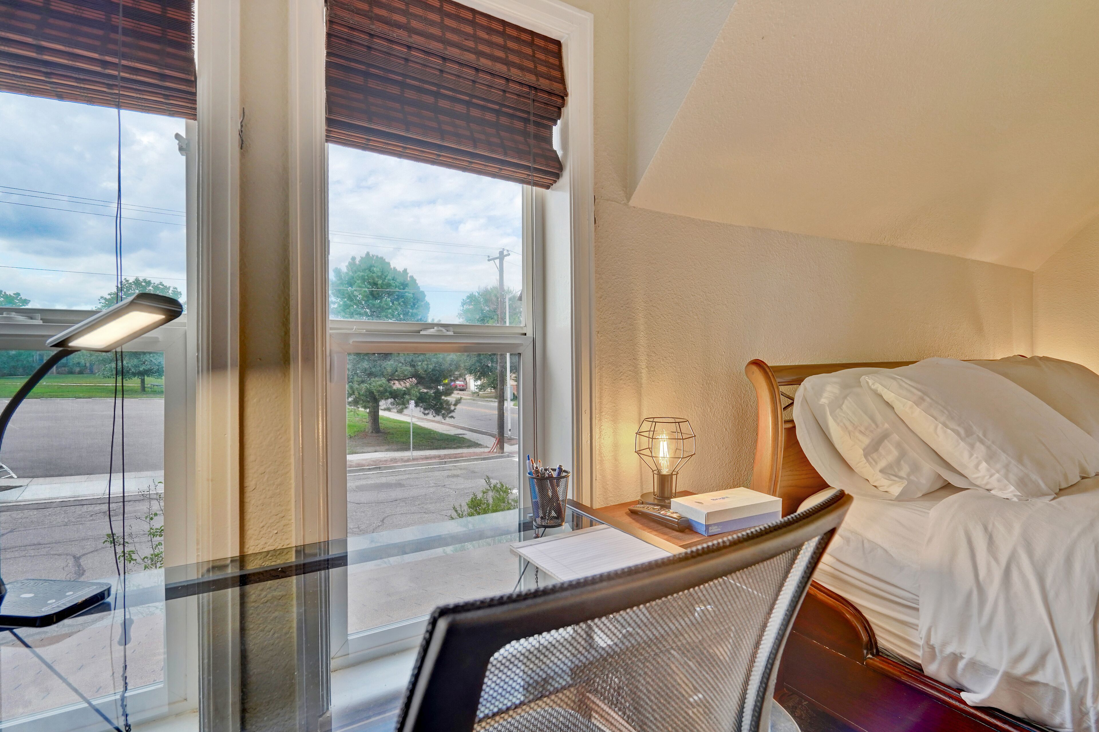 A hotel room with a bed, a desk with a lamp, and a view of the street through large windows.