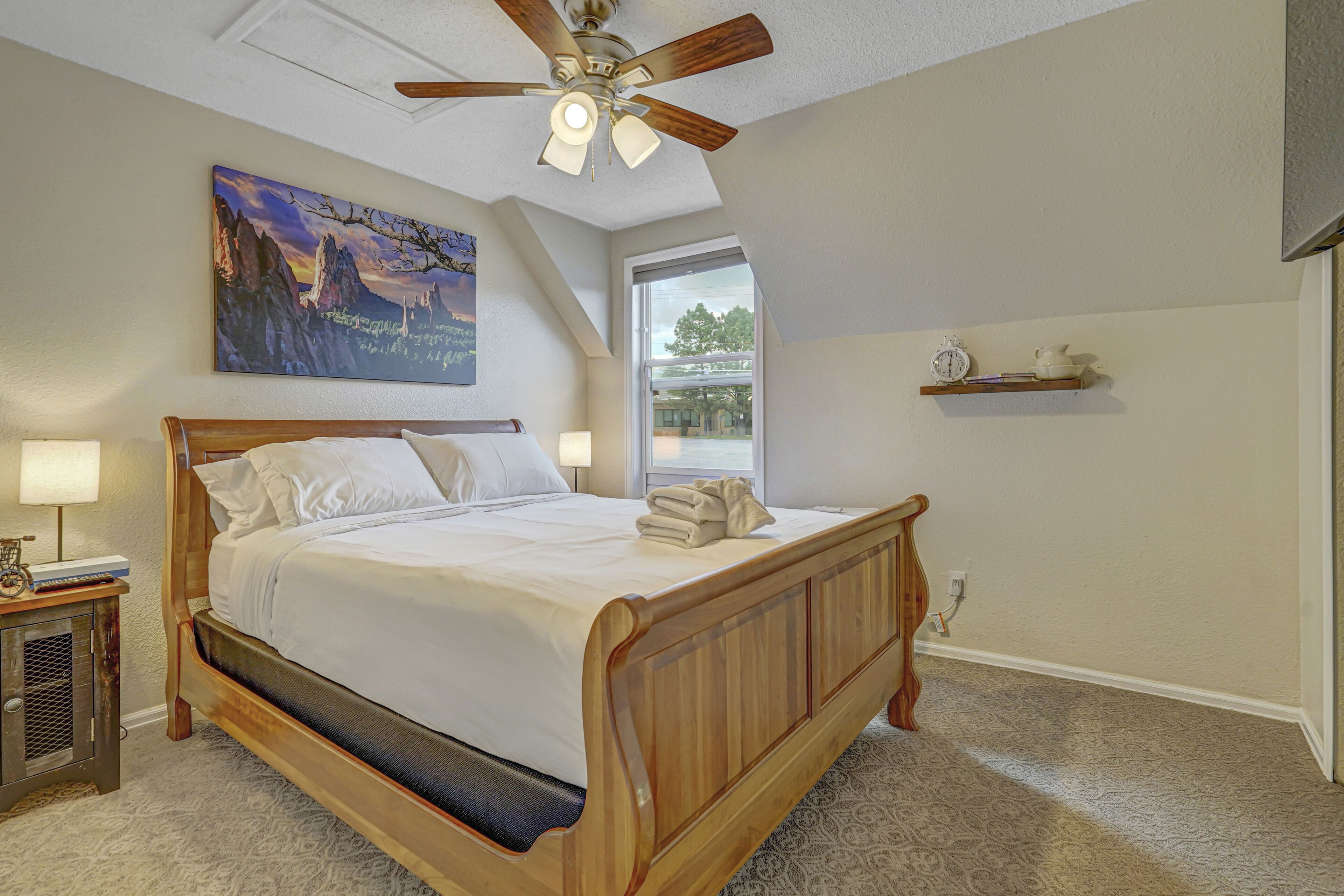A bedroom with a wooden bed, a ceiling fan, a painting, a window, and a shelf with books.