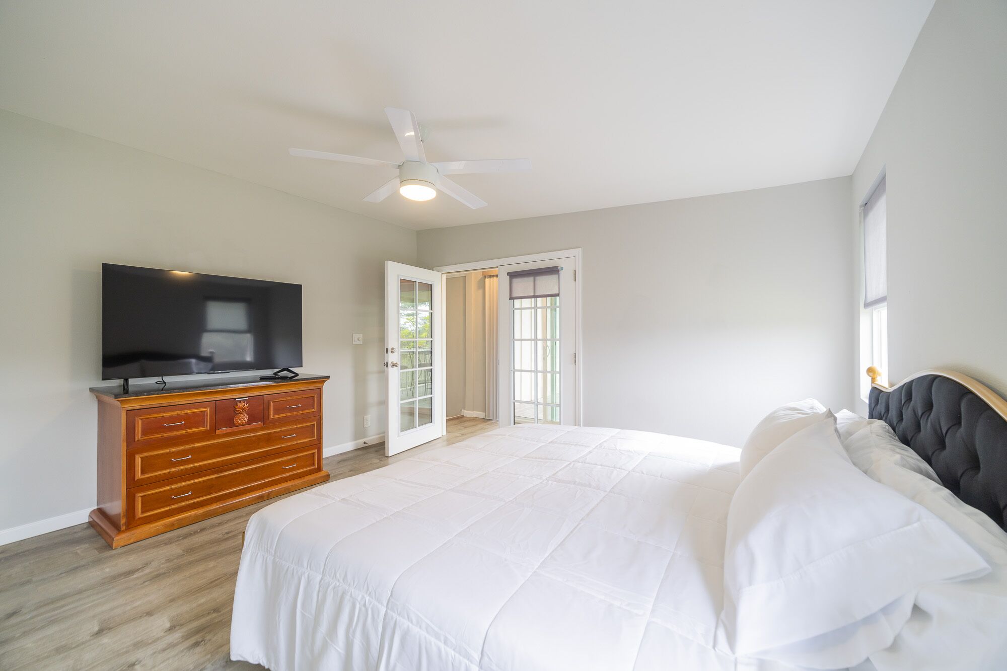 A bedroom with a bed, a TV on a wooden dresser, a ceiling fan, and a door leading to a balcony.