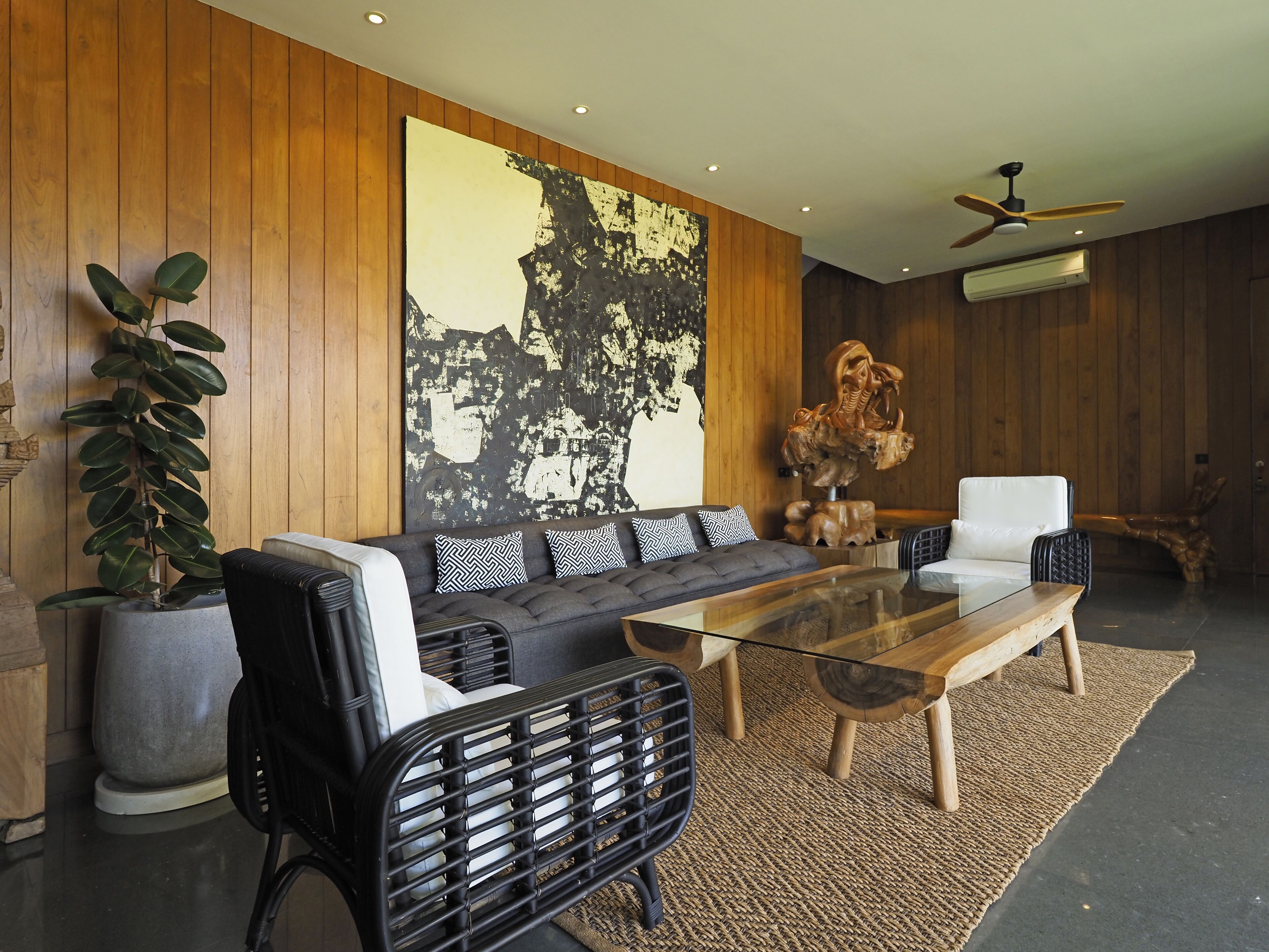 Un salon moderne avec un canapé, une table basse et des fauteuils. Un grand tableau abstrait orne le mur et une sculpture en bois est posée sur le sol.