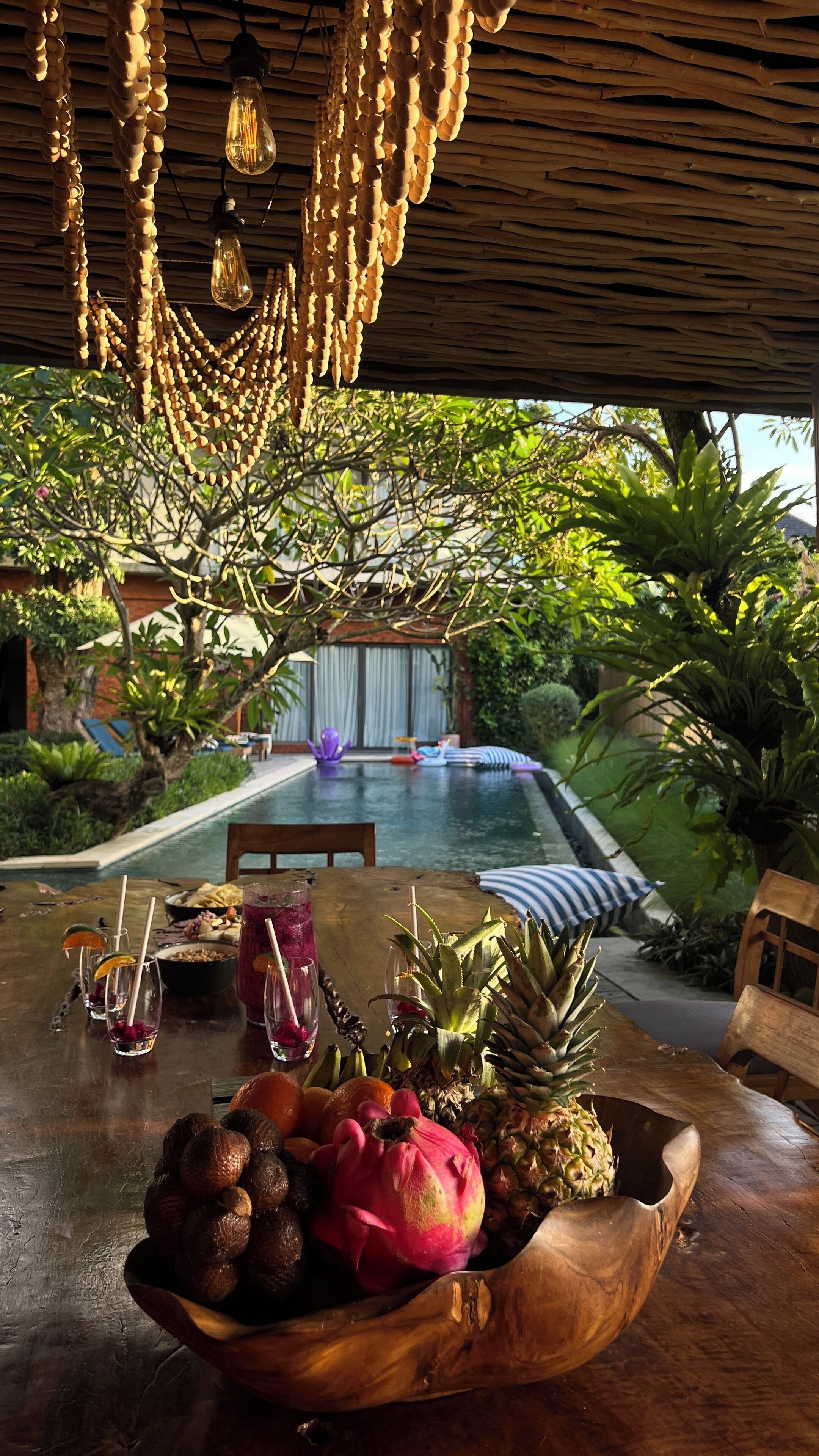 A wooden table with a bowl of fruit, a drink, and a poolside setting with a view of a building and greenery.