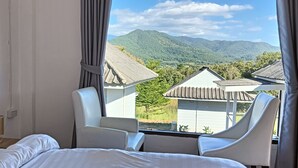 Une chambre d’hôtel avec un lit, deux chaises blanches et une fenêtre donnant sur un paysage de montagnes et de bâtiments.