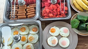 Un buffet proposant une variété d’aliments, notamment des hot-dogs, de la pastèque, des œufs au plat, des bananes et des légumes verts à feuilles.